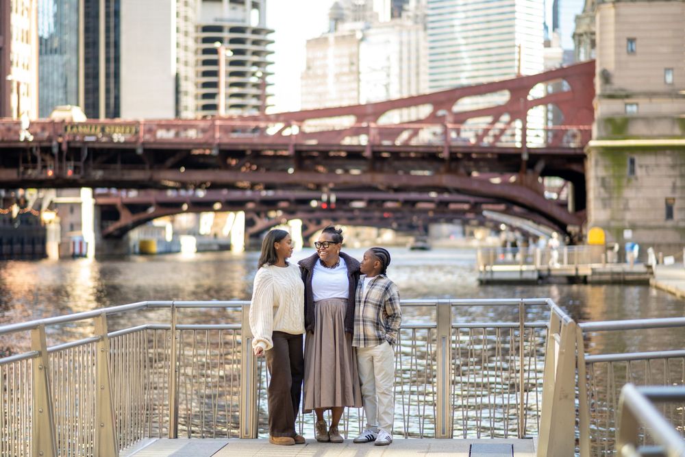 Three women stand together on a riverside walkway with a red bridge and city skyline in the background.