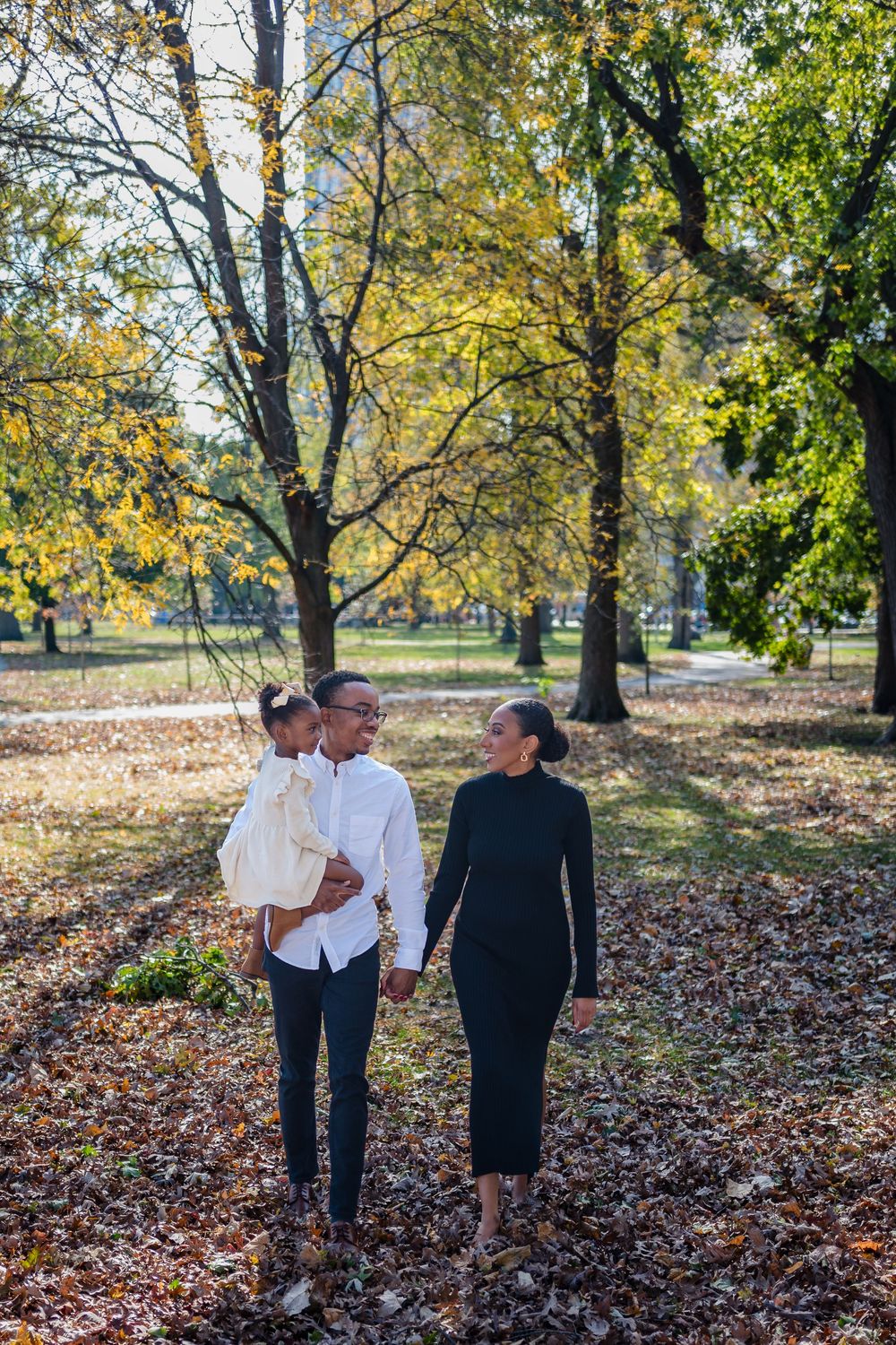 Two people walk together through a scenic autumn park with fallen leaves covering the ground on a sunny day.