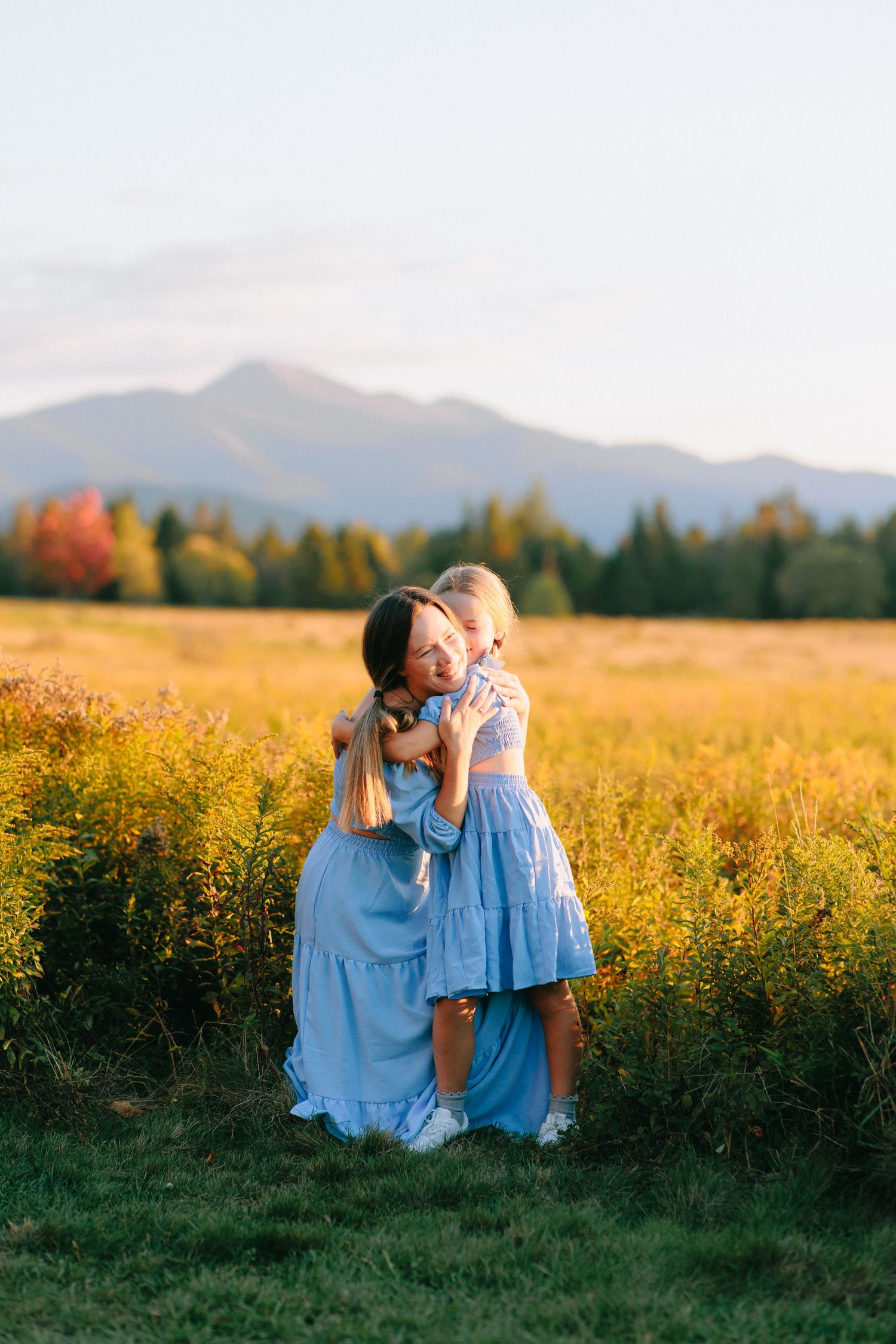 People in blue dresses share tender moments in a golden field with mountains in the background at sunset.