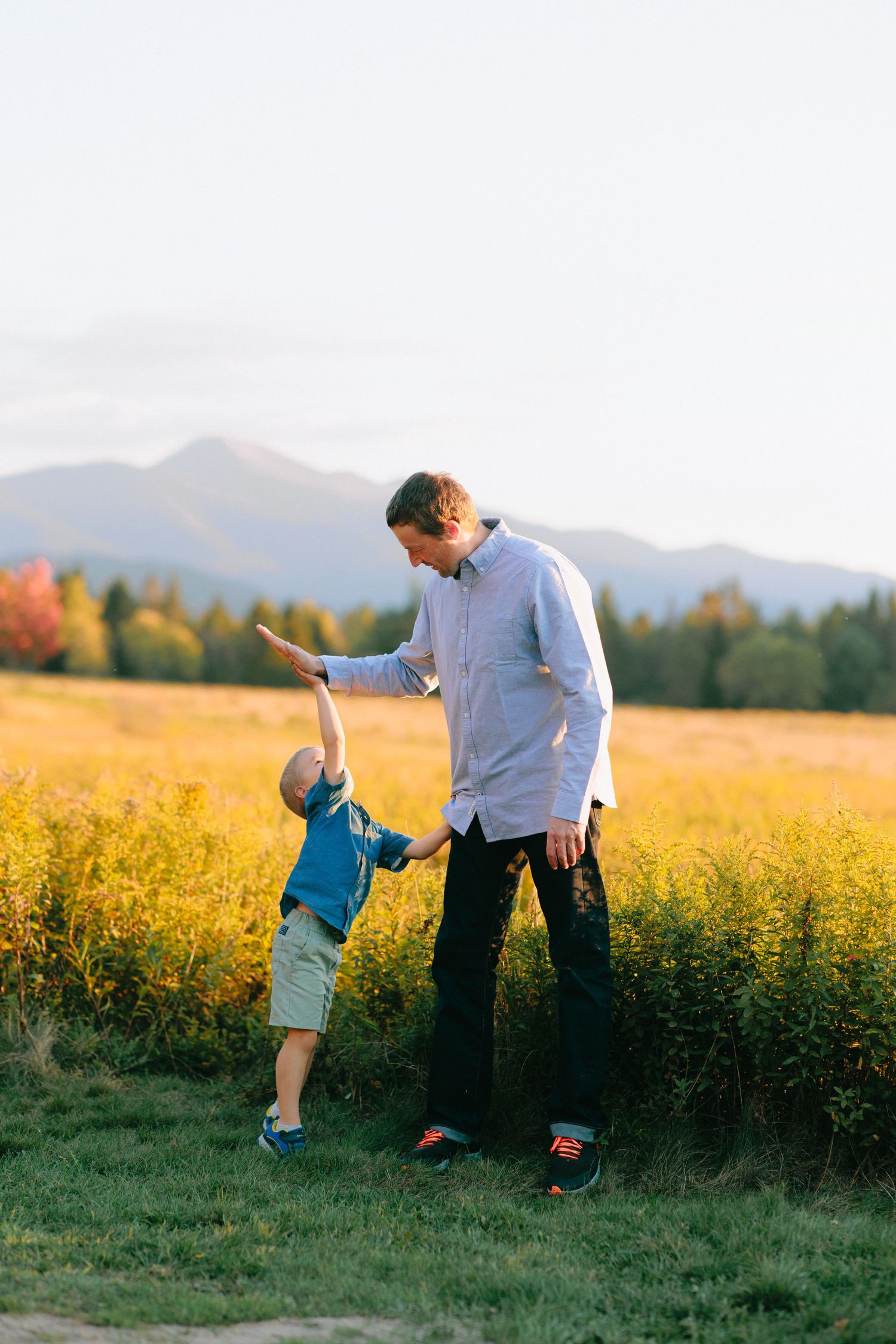 A parent and child play together in a sunny meadow with mountains visible in the distance.