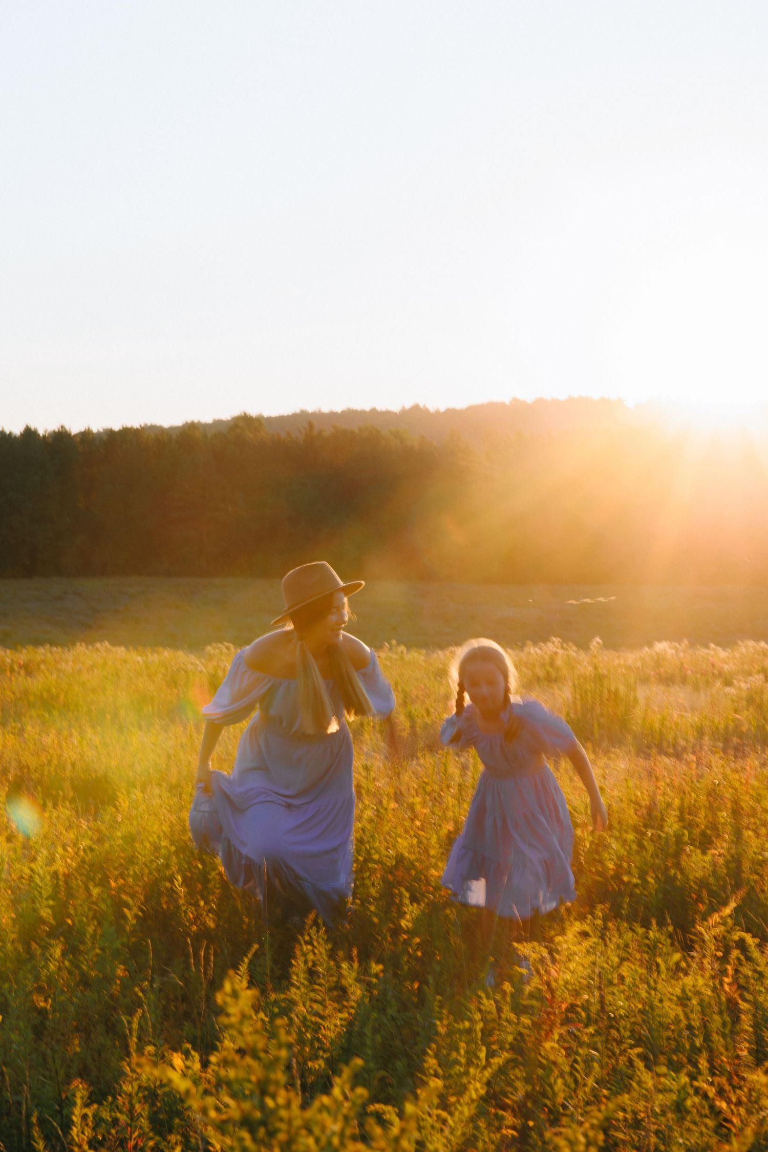 Two figures sit together in a golden meadow at sunset as warm light bathes the wildflowers and grass.