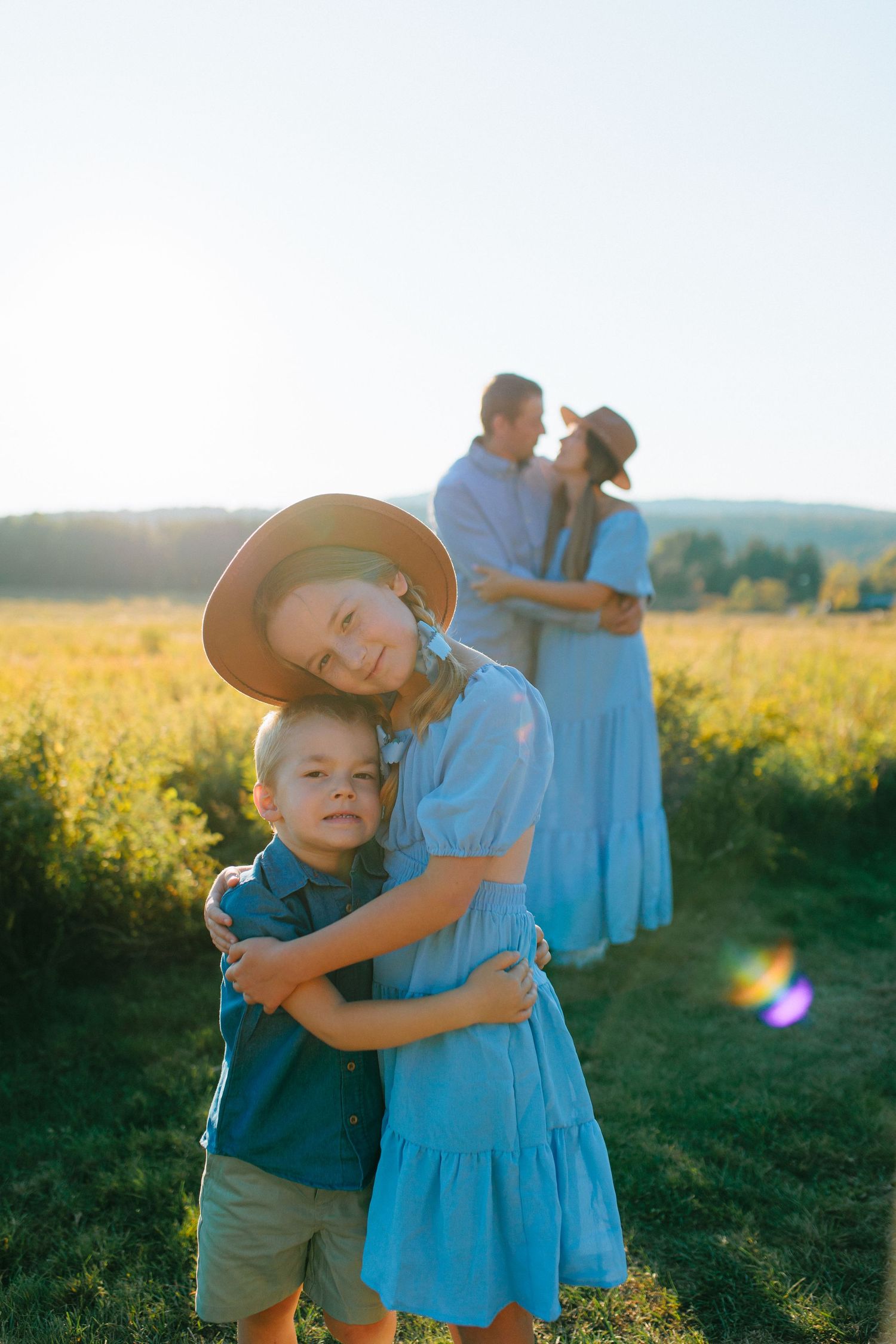 Two children share a sweet embrace in a sunlit meadow of yellow wildflowers at golden hour.