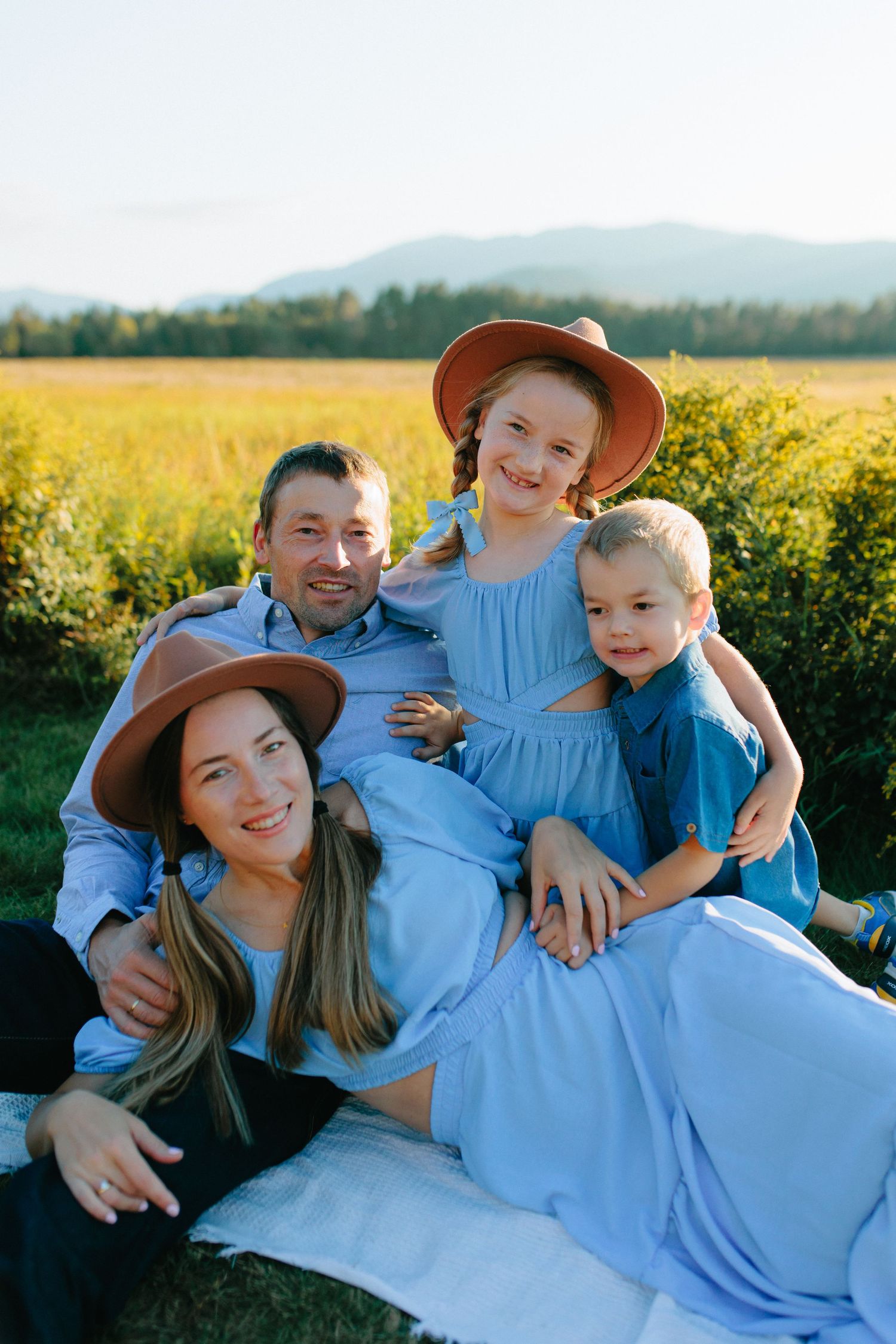 Happy family relaxing together in a scenic field during golden hour wearing blue and brown outfits.
