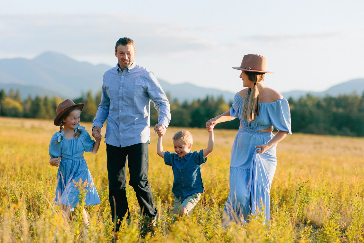 Family in denim outfits walking together through a golden wheat field at sunset with mountains in background.