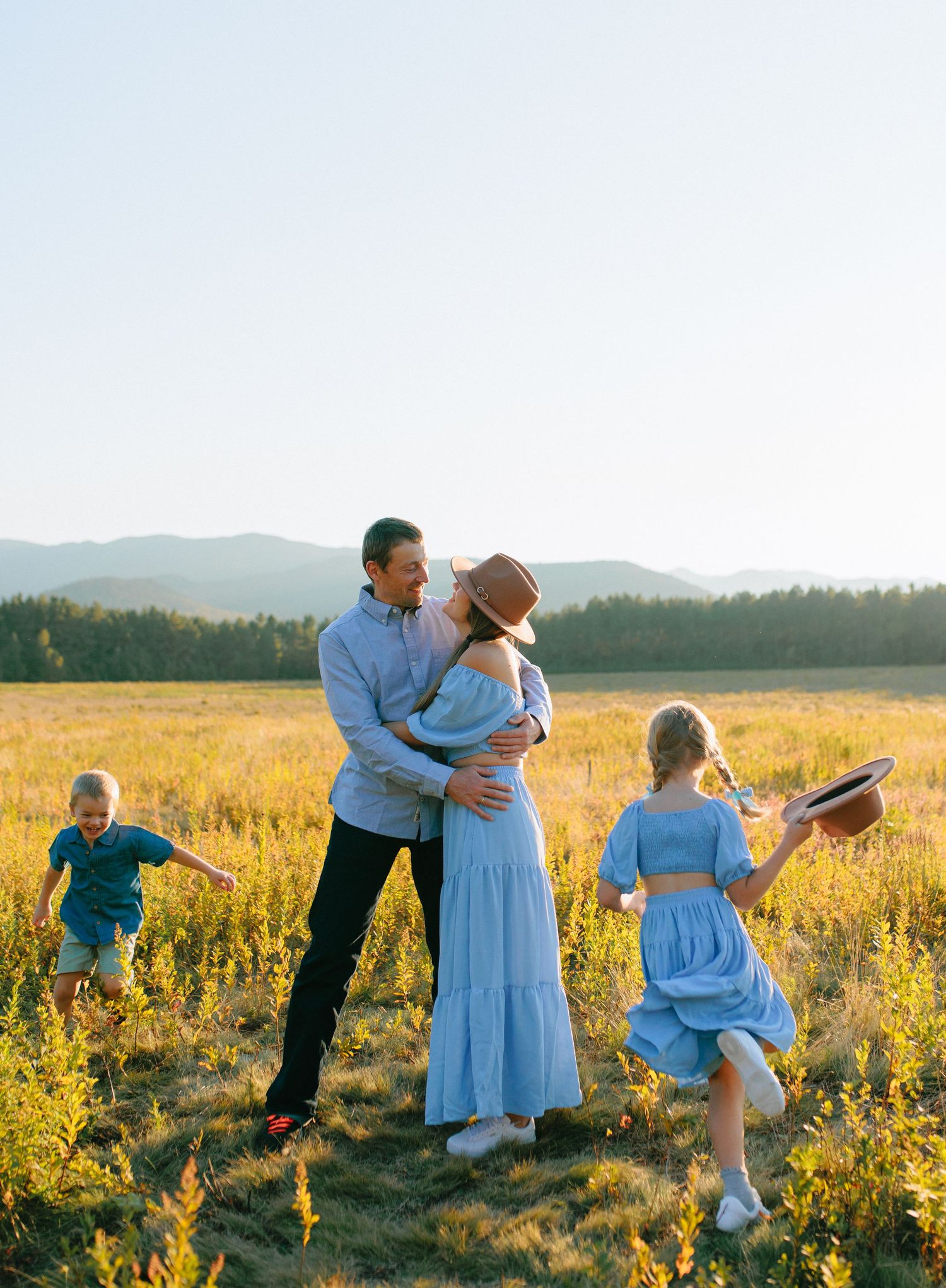 A family in blue dresses and casual wear dances together in a golden sunlit field during sunset.