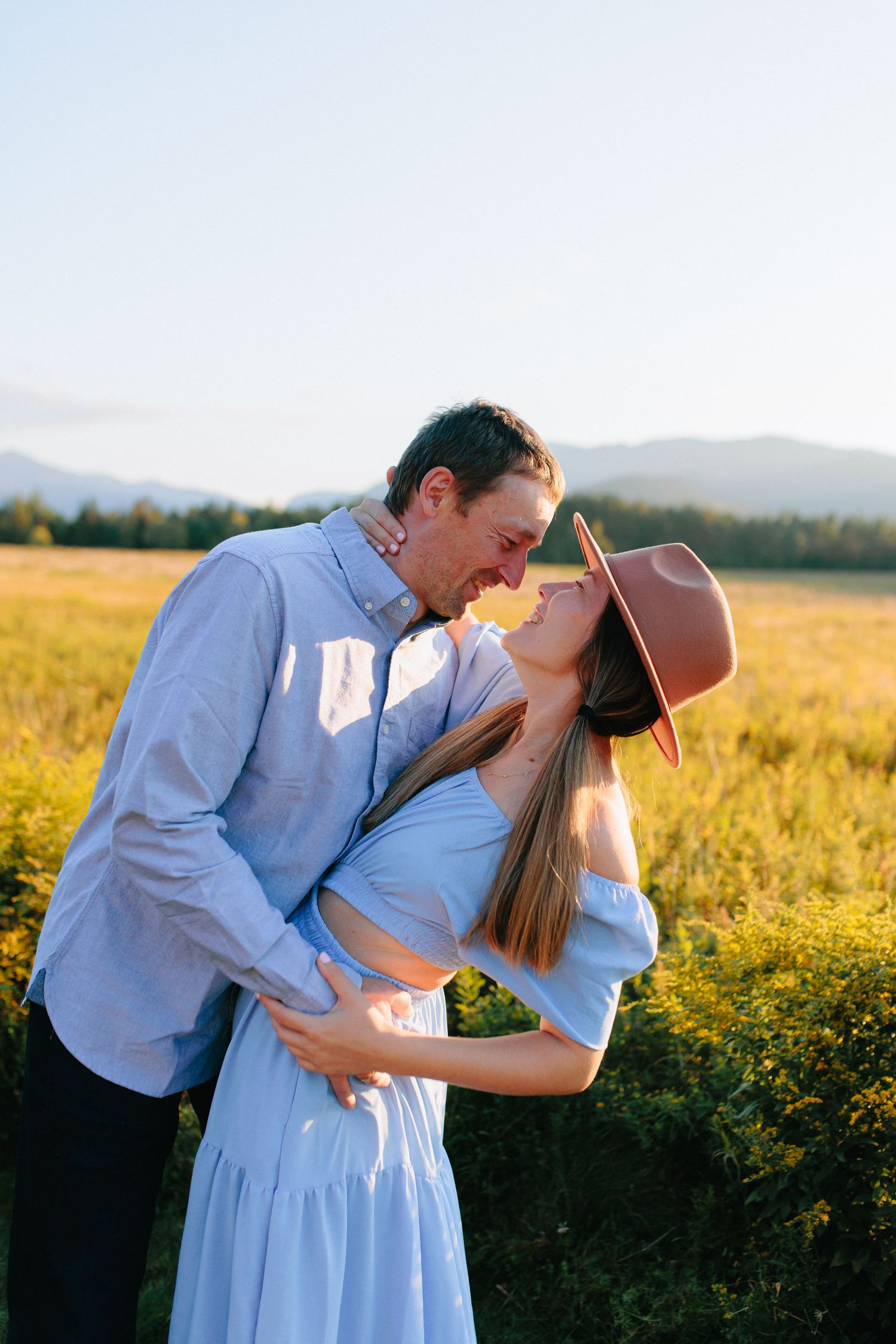 A romantic couple embraces in a golden field at sunset with mountains in the background.