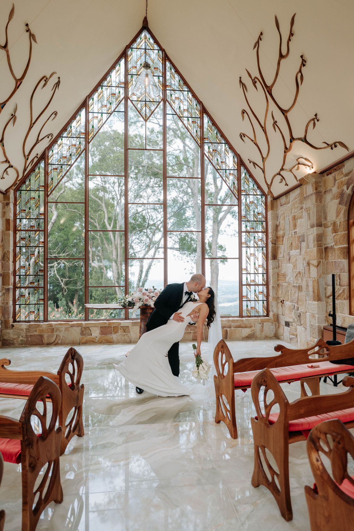 Panoramic view of a modern chapel interior with stone walls, wooden pews, and geometric windows overlooking nature.