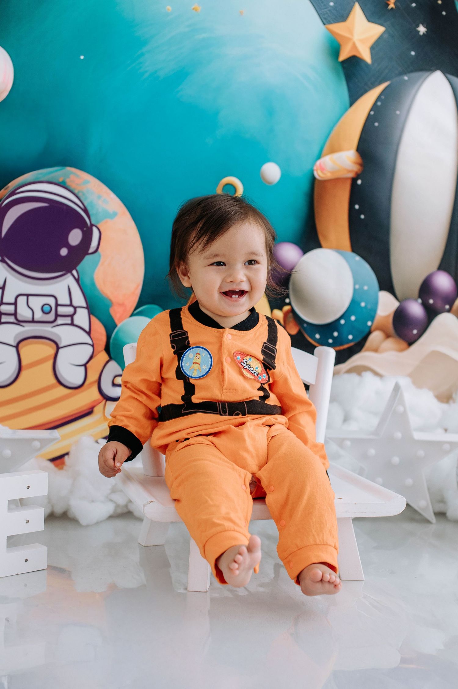 Baby in orange astronaut costume sits among space-themed decorations with stars and moons against turquoise backdrop.