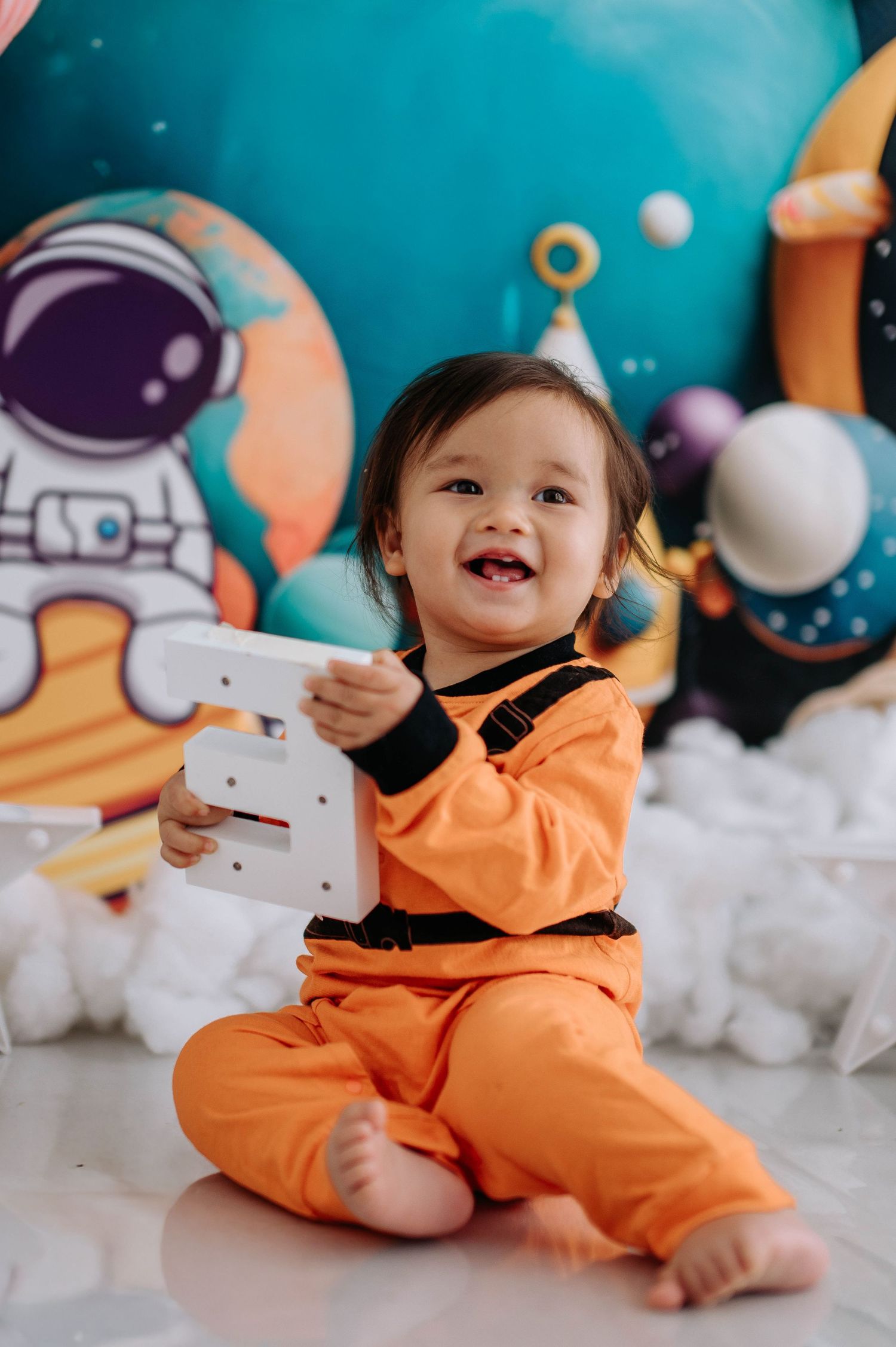 A small child in an orange astronaut costume plays against a space-themed backdrop with white cotton clouds.