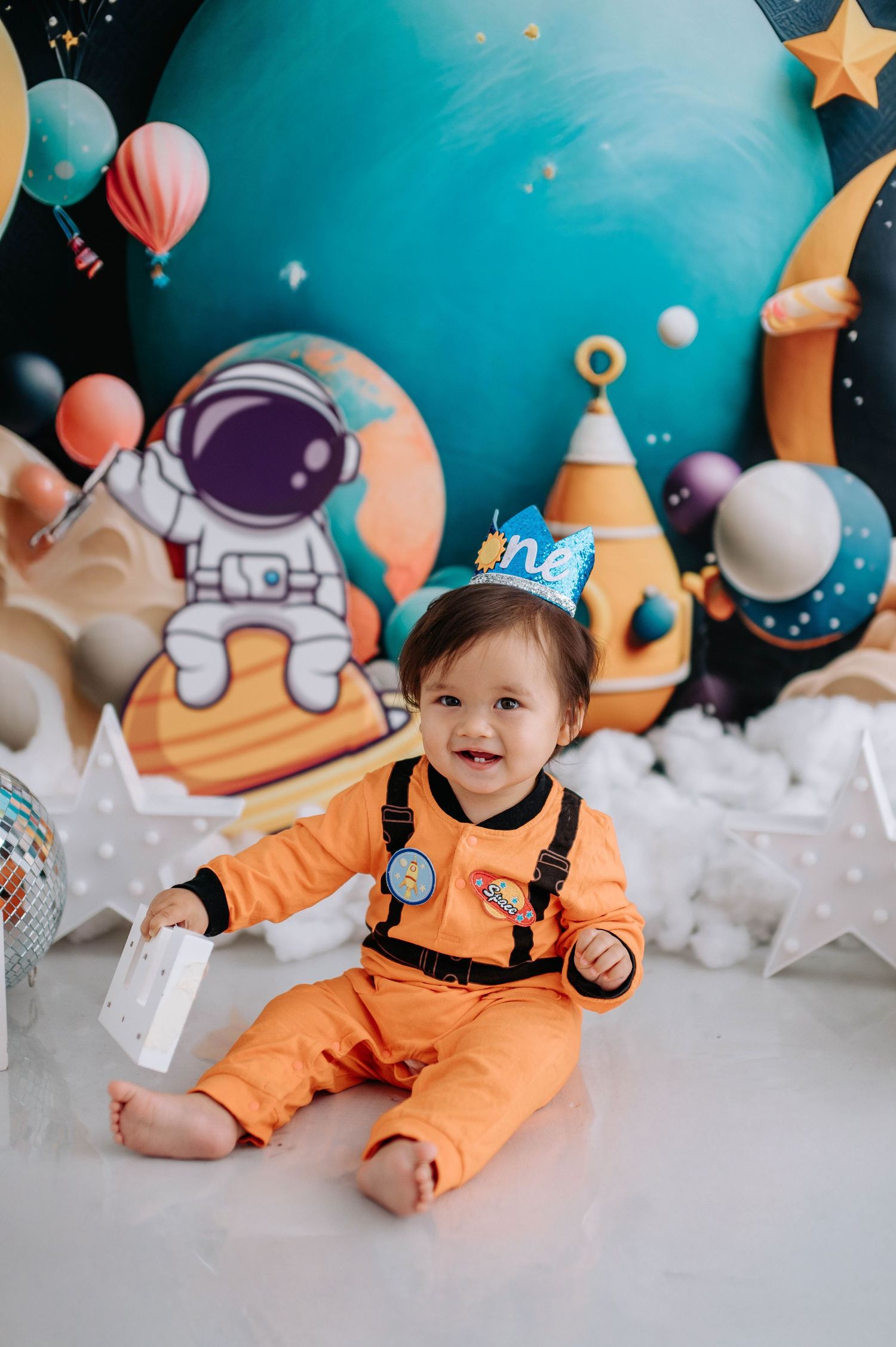 Adorable space-themed photo setup with orange astronaut outfit against a turquoise backdrop with stars and planets.