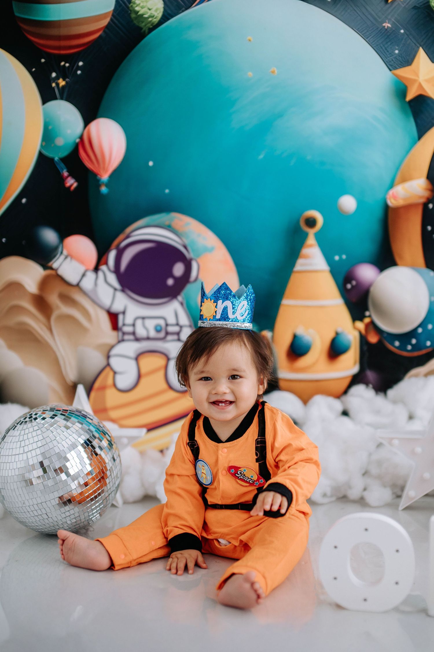 Cute space-themed photoshoot featuring orange spacesuit costume with silver disco ball and celestial decorations.
