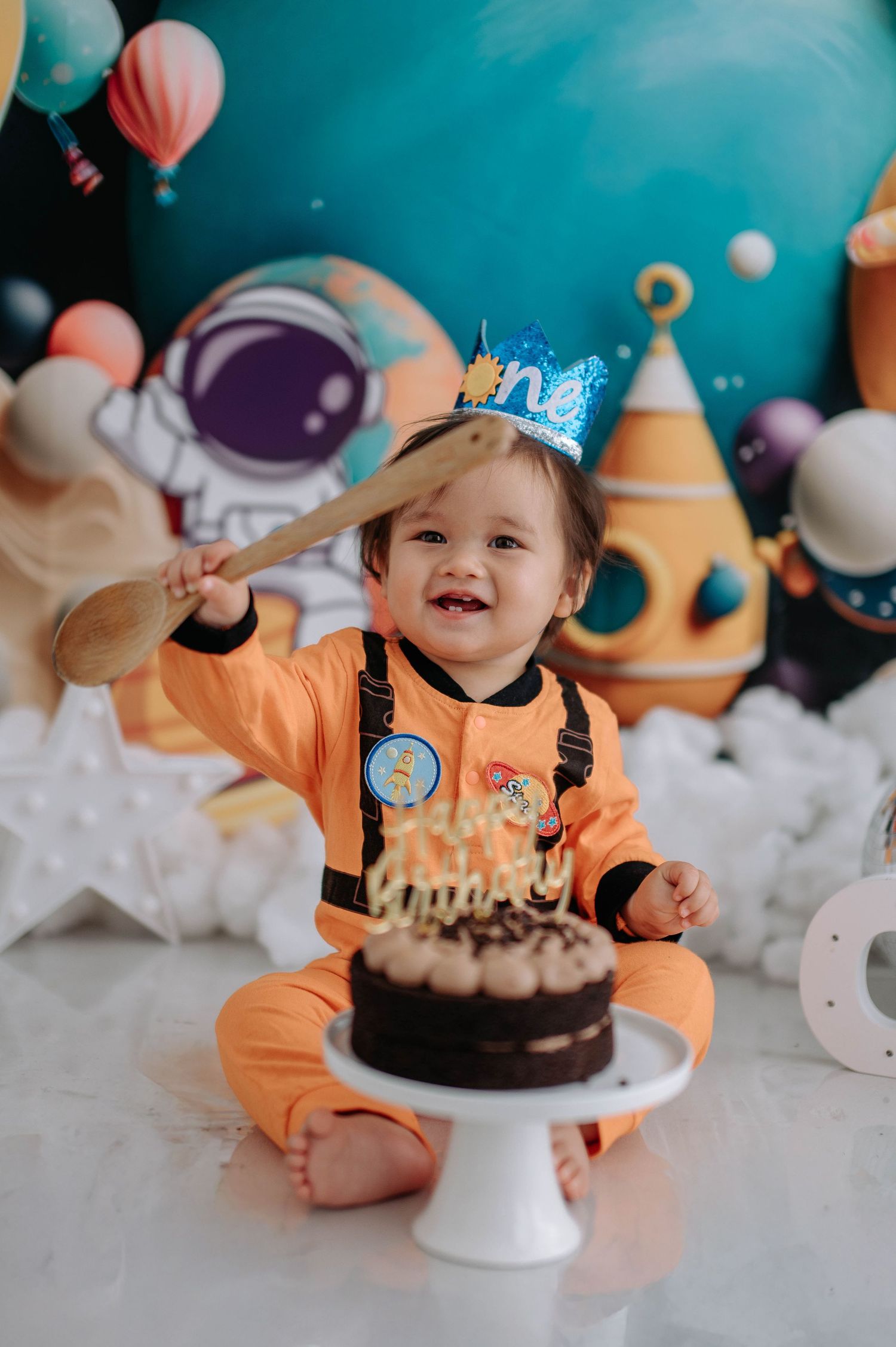 Adorable cake smash photo session with orange outfit and chocolate cake against blue backdrop and party decorations.