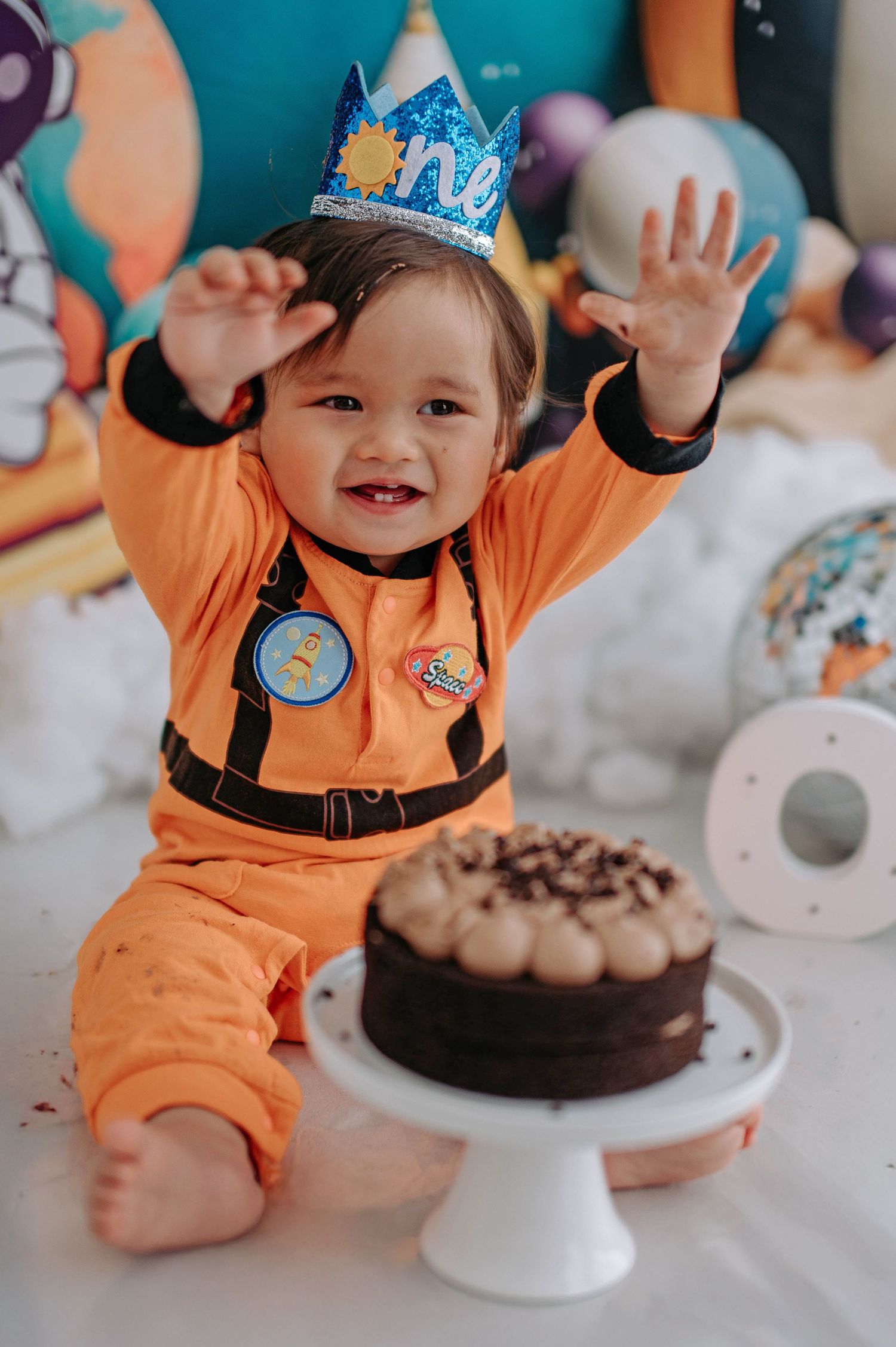 A baby in an orange outfit celebrates their first birthday with a chocolate cake on a white cake stand.