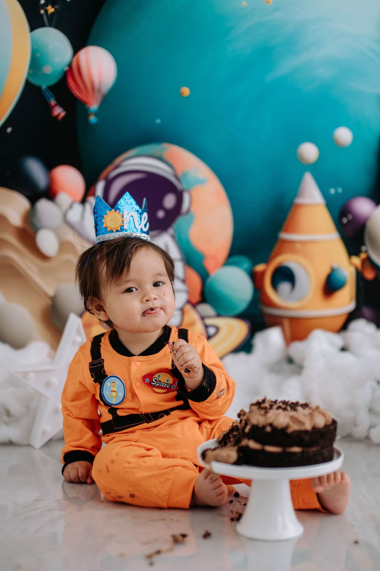 A small child in an orange astronaut costume poses with a chocolate cake during a space-themed birthday celebration.