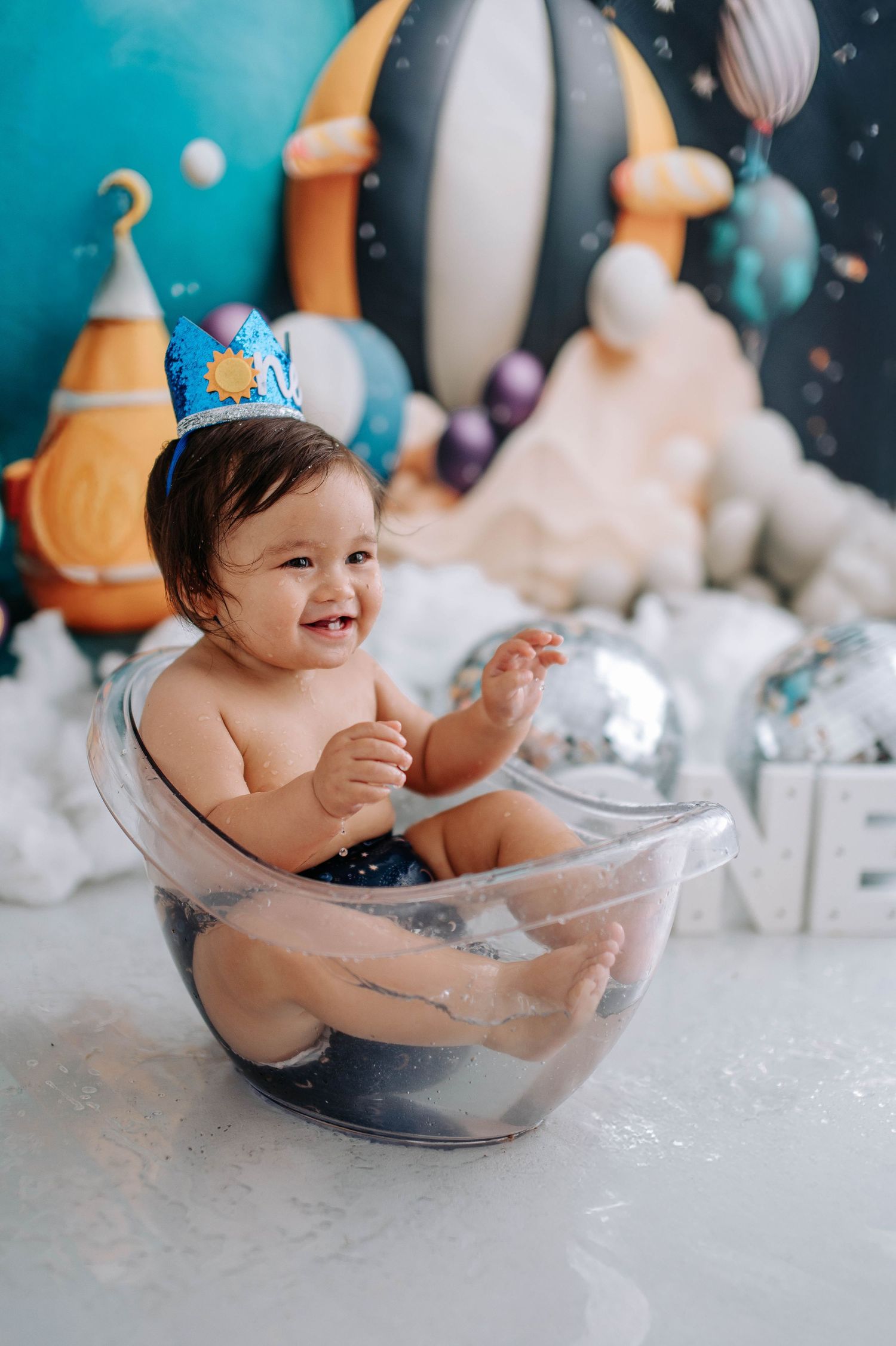 Baby enjoying bath time in clear tub with festive decorations and blue background.