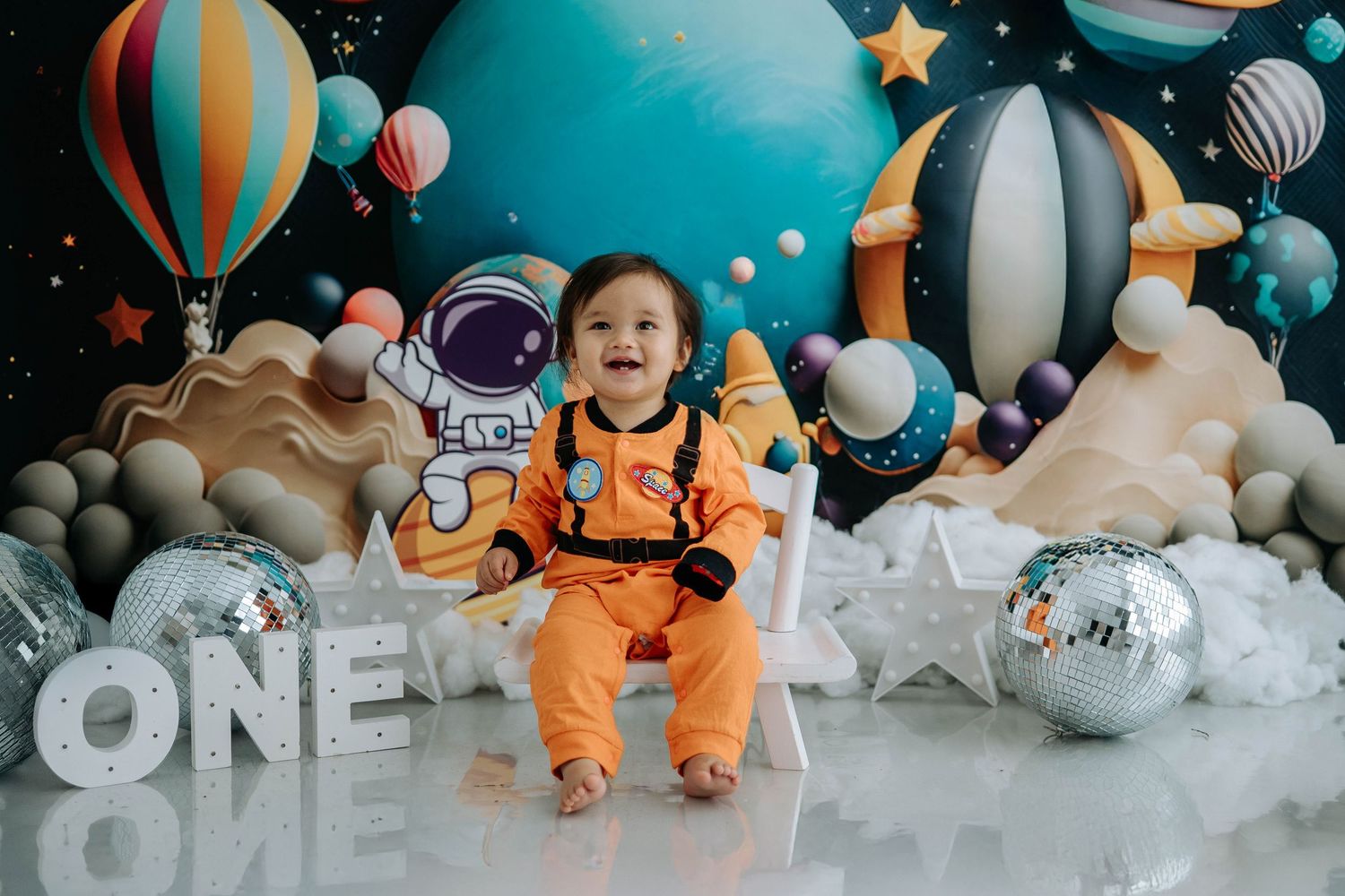 Astronaut-themed first birthday party setup with hot air balloons, clouds, and orange space suits against a blue backdrop.