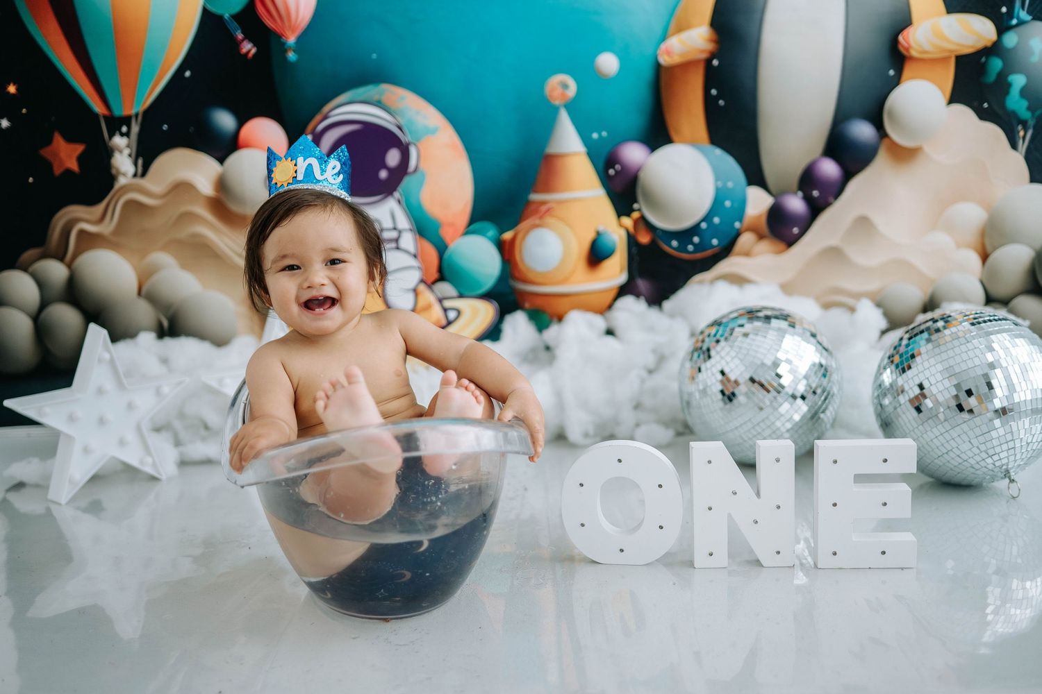 First birthday celebration setup with silver disco balls, colorful balloons, and clouds against a dark background.
