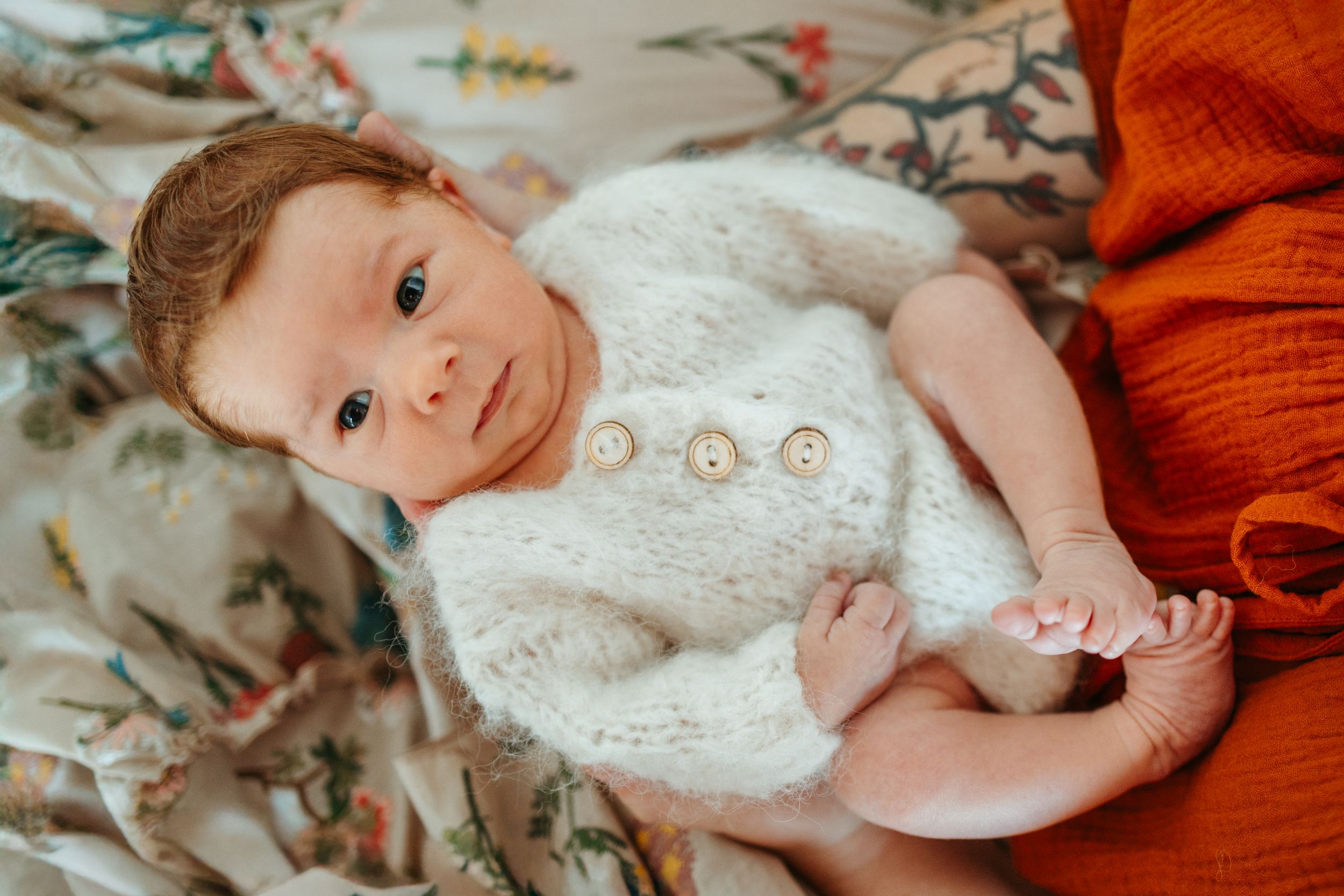 A baby in a white knit sweater lies on a floral patterned blanket while being held by someone in orange clothing.