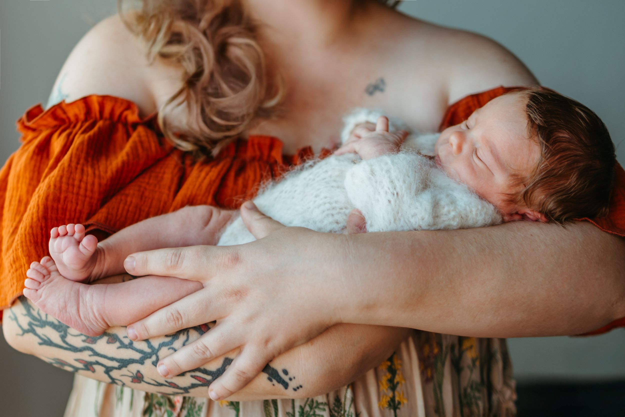 Hands gently cradle a sleeping newborn baby wrapped in white swaddling cloth against a soft gray background.