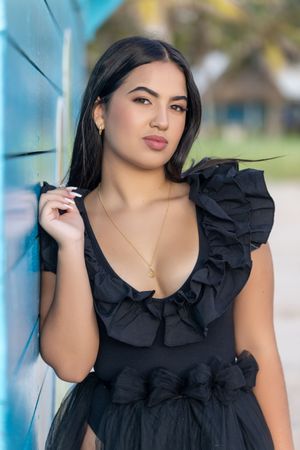 Young woman in black ruffled dress posing against a blue wall outdoors.
