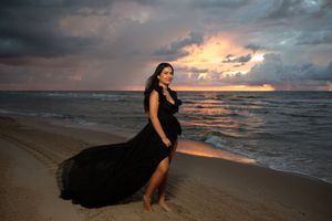 Pregnant woman in flowing black gown poses on sandy beach at dramatic sunset with stormy skies and ocean waves.