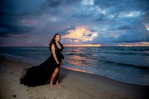 Woman in flowing black gown poses on beach at dramatic sunset with stormy skies and ocean waves.
