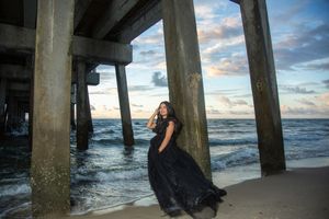 Woman in black gown posing under a pier on the beach at sunset with dramatic cloudy skies.