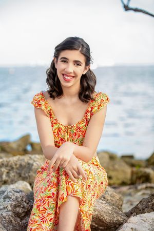 Young woman in colorful floral dress sitting on rocks by the ocean, smiling outdoors.