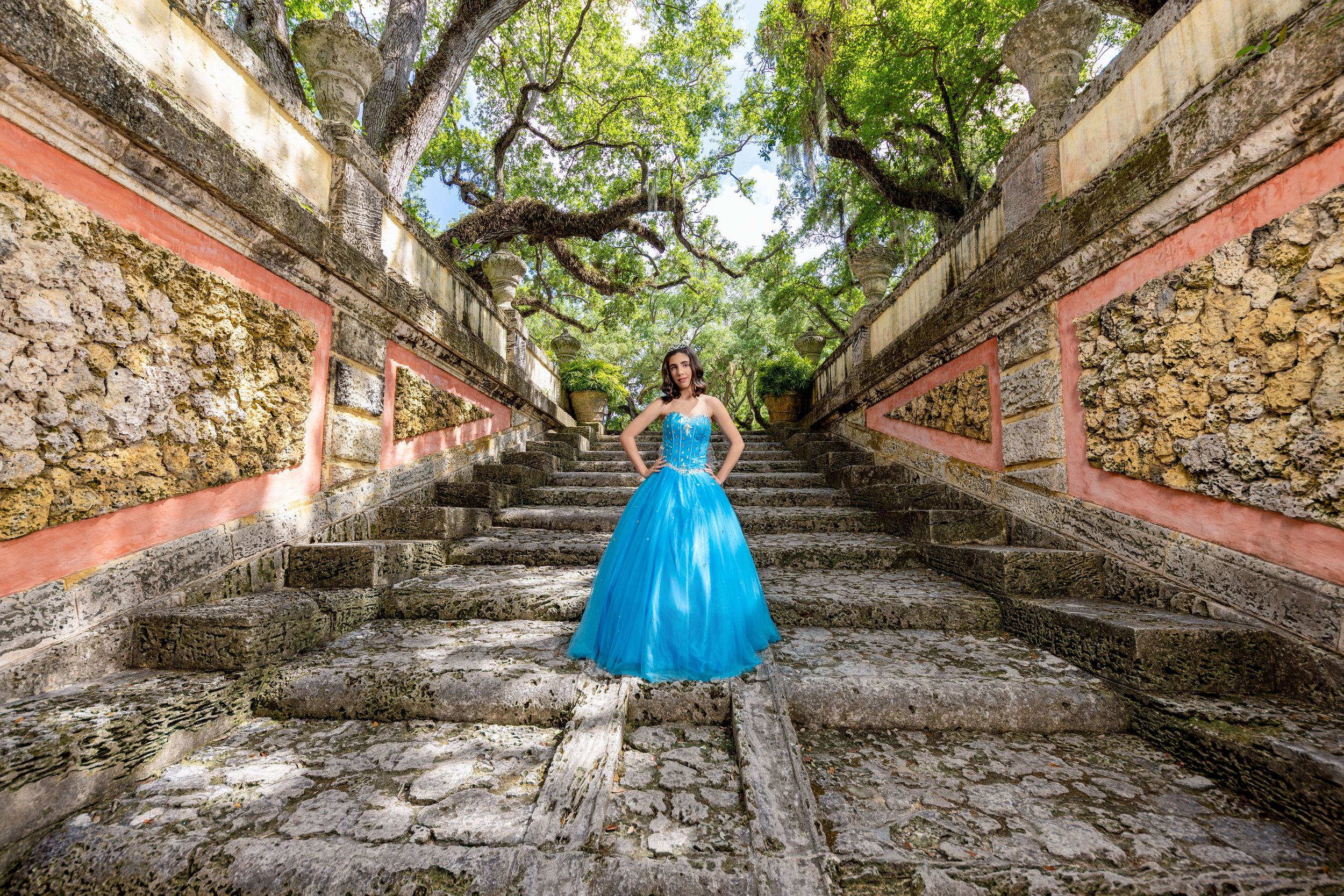 Young woman in a blue ball gown stands on ancient stone steps surrounded by lush trees and rustic walls.