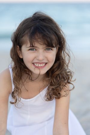 Smiling young girl with curly brown hair wearing a white dress at the beach.