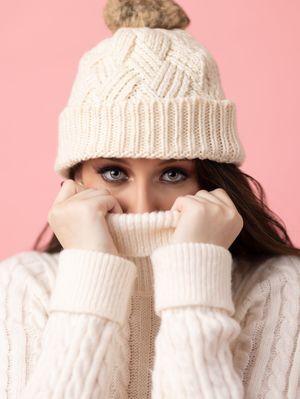 Woman in cream knit beanie hat and turtleneck sweater on pink background.