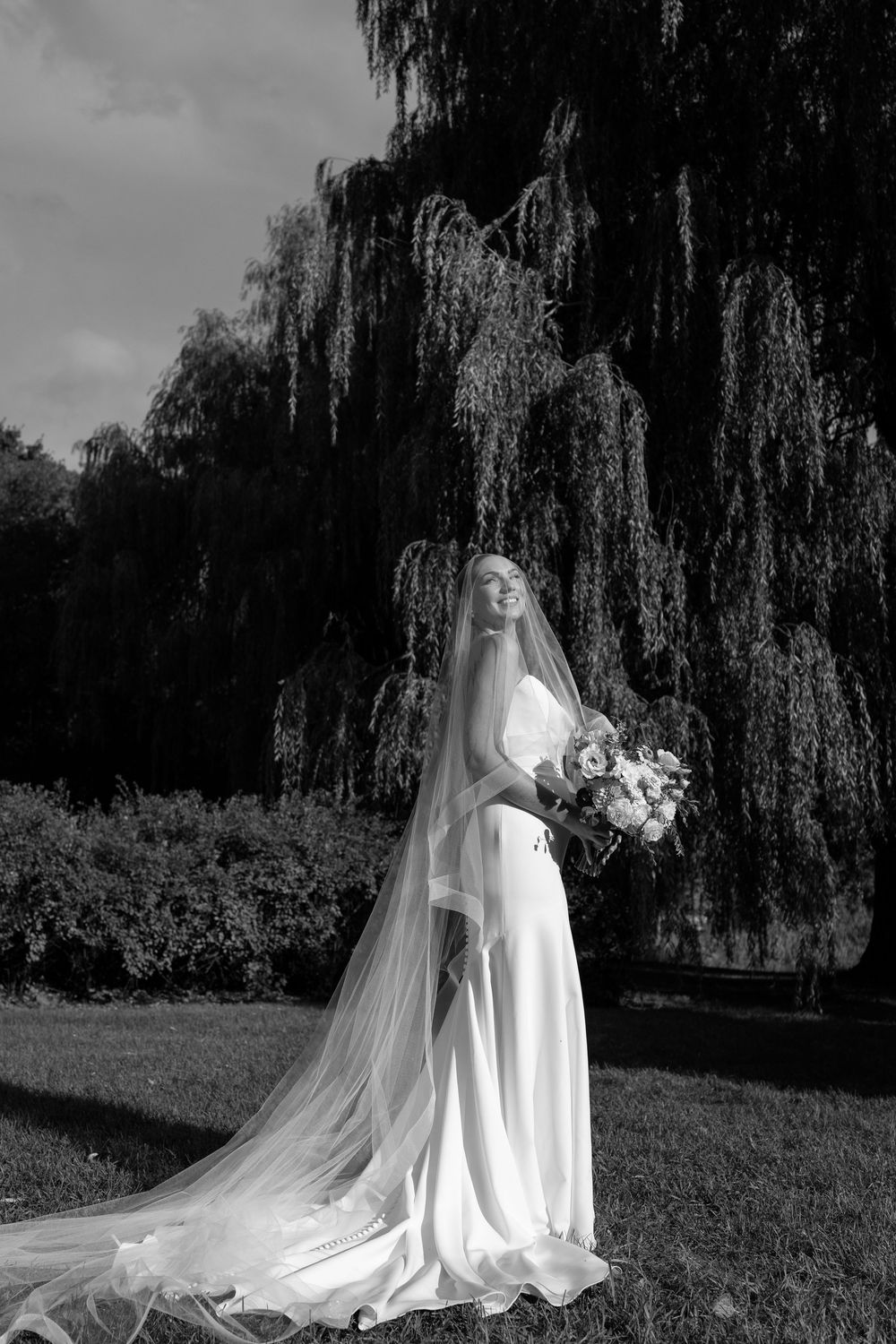 Black and white wedding photo series showing a bride in flowing gown and veil posing under dramatic weeping willow trees.