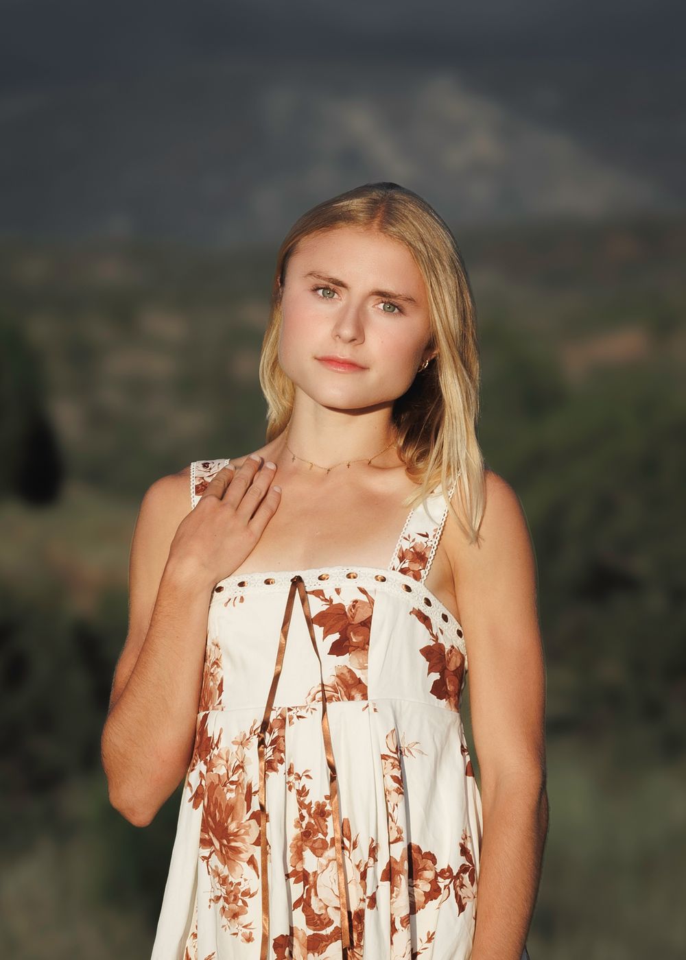Young blonde woman in white floral sundress posing thoughtfully with mountain landscape at golden hour.