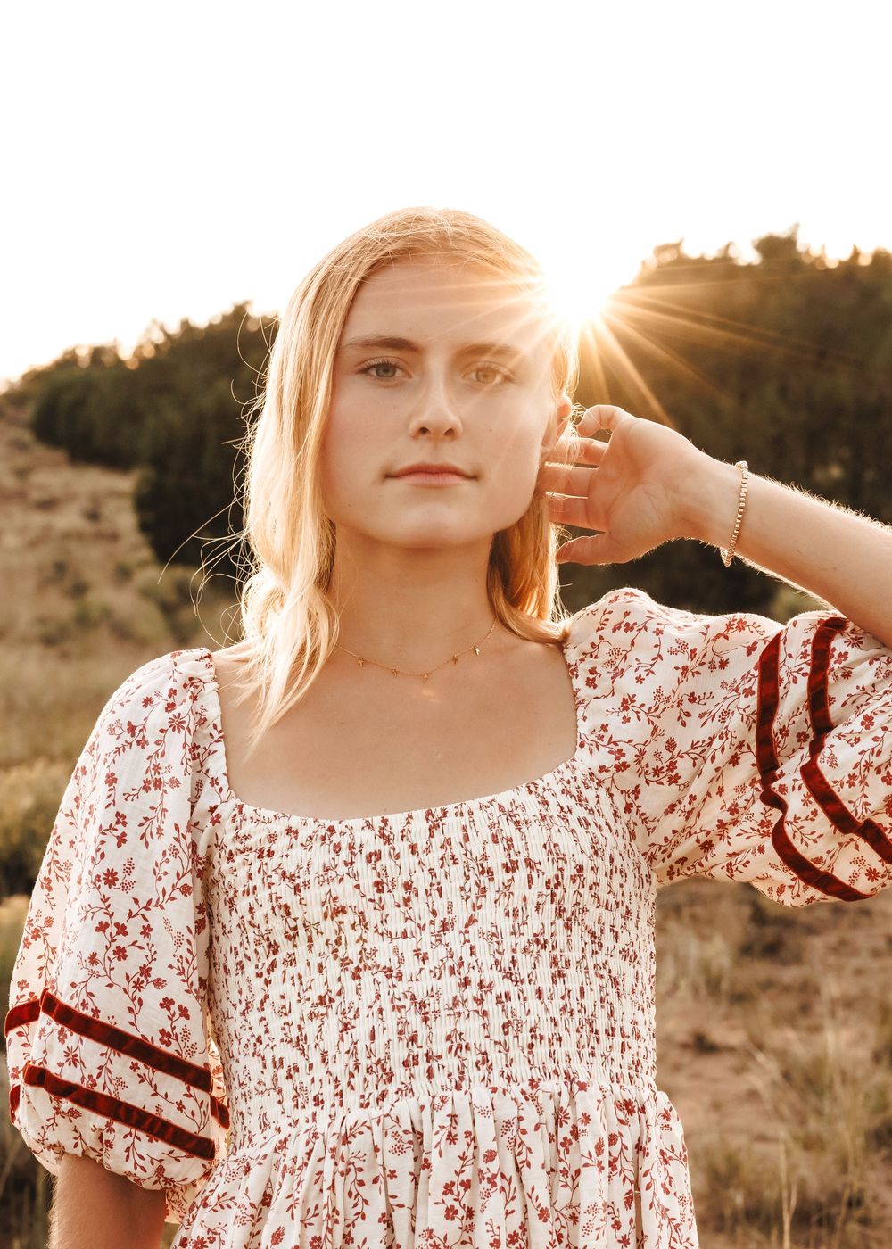 Blonde teen in white floral smocked dress with sun flare behind her in a natural outdoor setting.