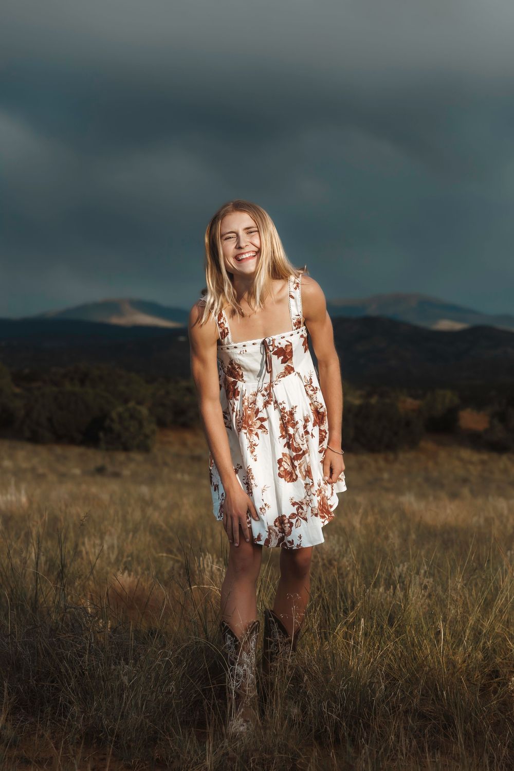 Santa Fe senior teen in white floral sundress and cowboy boots in golden field with stormy New Mexico sky.