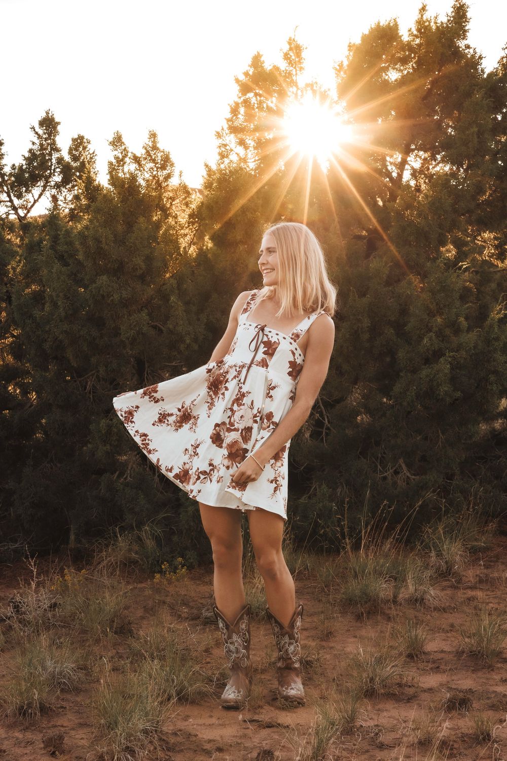 Blonde teen in white floral sundress and cowboy boots smiling at sunset with golden sun flare.