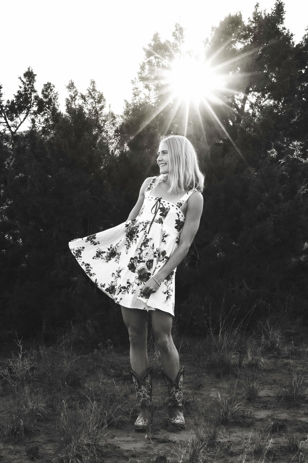 Black and white photo of smiling blonde teen in floral dress with sun flare through trees.