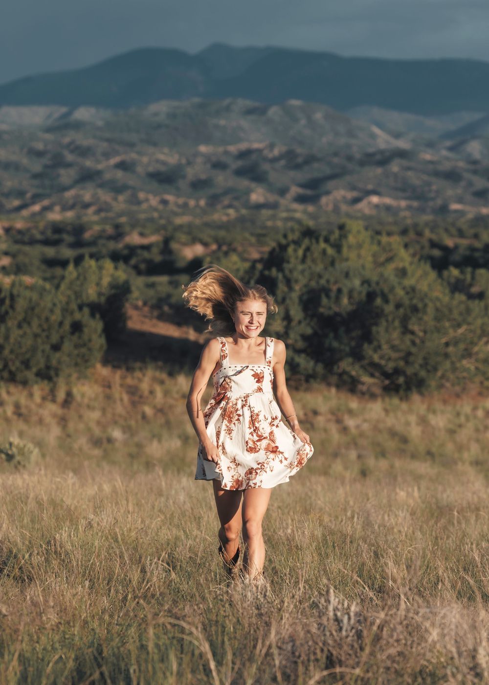 Blonde teen in floral sundress running through golden meadow with dramatic mountain range in background.