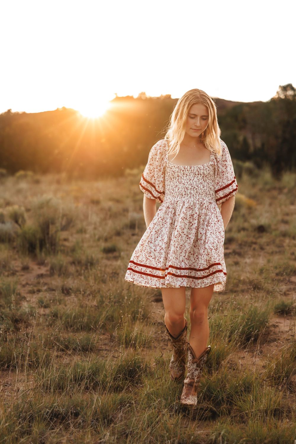 Santa Fe senior in floral mini dress and cowboy boots walking in golden sunset field.
