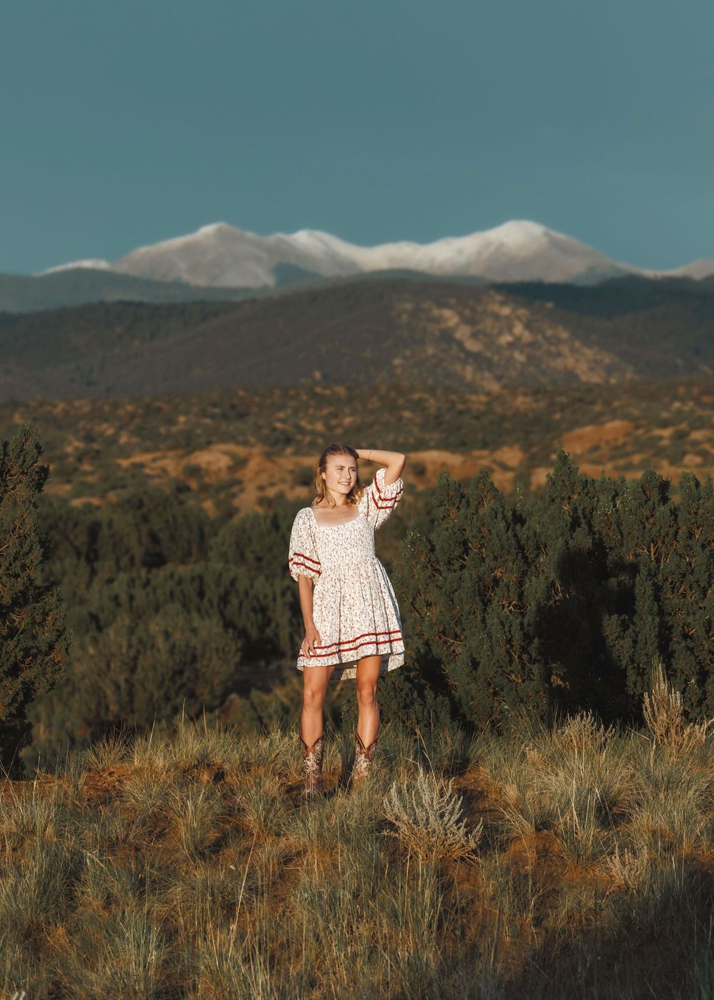 Smiling blonde woman in floral dress standing in field with snow-capped mountains behind.