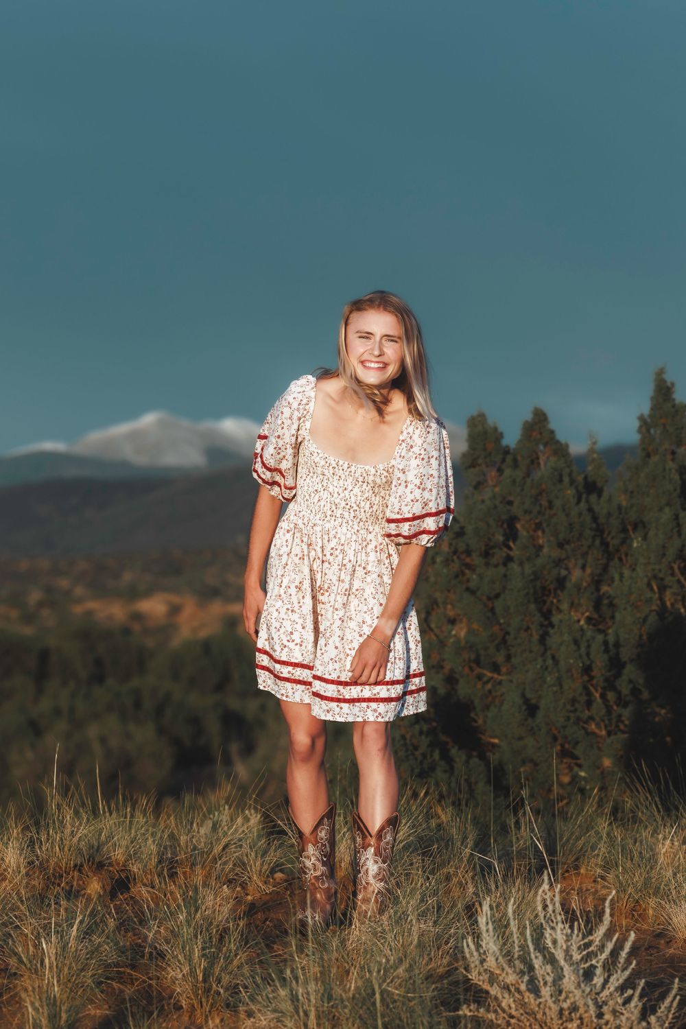 Smiling blonde woman in floral dress and cowboy boots standing in field with mountains.
