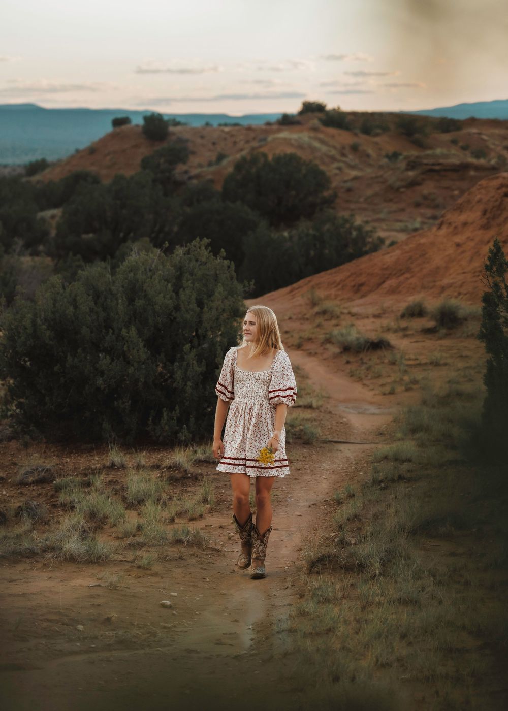 Blonde woman in floral dress and cowboy boots walking on dusty desert trail at dusk.