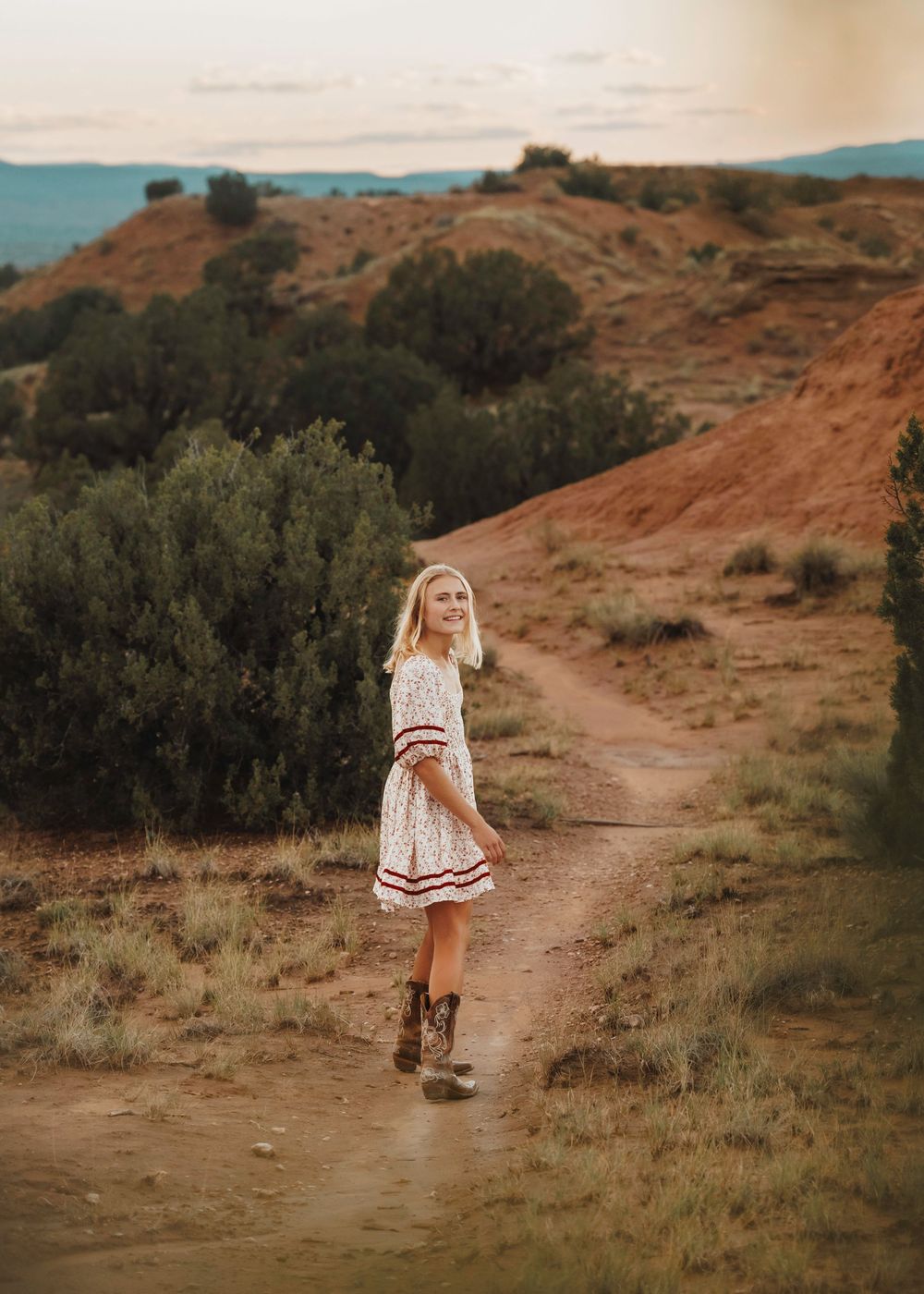 Blonde woman in floral dress and cowboy boots smiling on desert dirt path at sunset.