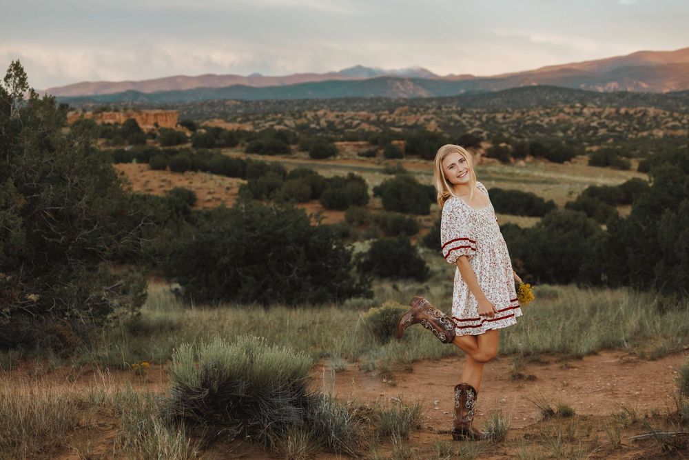 Blonde woman in floral dress holding flowers on scenic desert hilltop at golden hour.