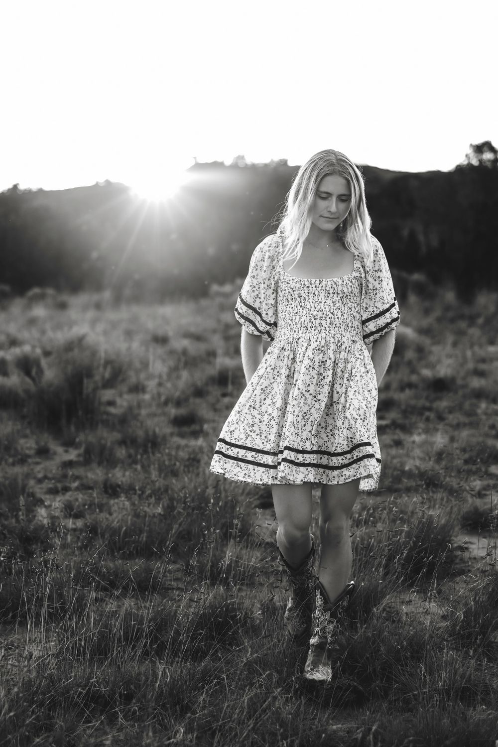 Black and white photo of Santa Fe senior in floral dress walking through field with sunburst behind her.