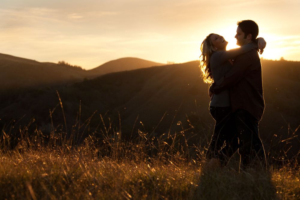 Two silhouettes embrace in a golden field at sunset with rolling hills in the background.