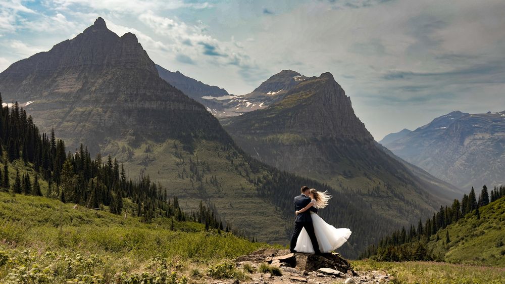 A couple embraces on a grassy mountain slope with dramatic peaks of Glacier National Park towering in the background.