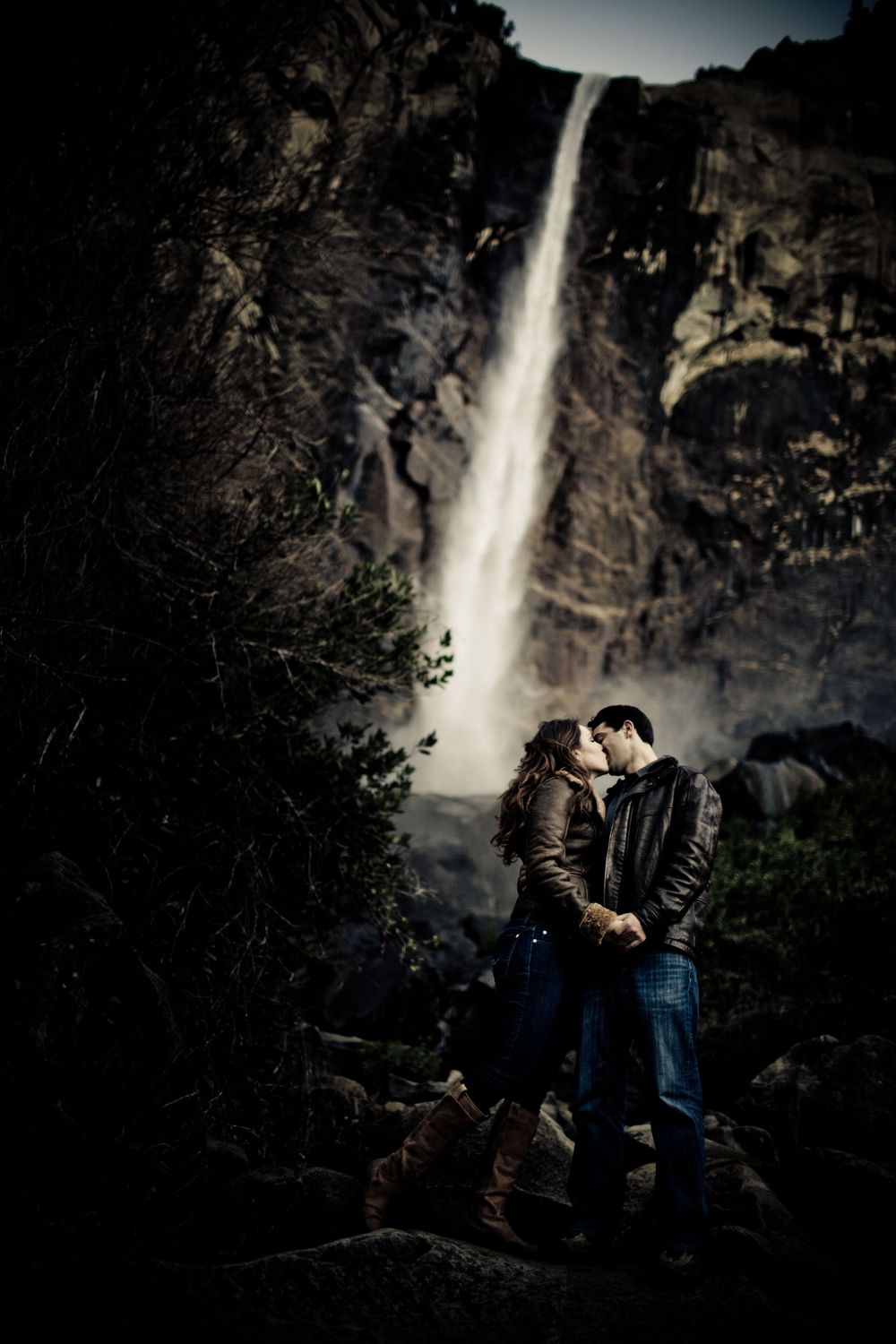 A couple shares an intimate moment beneath a dramatic waterfall cascading down rocky cliffs.