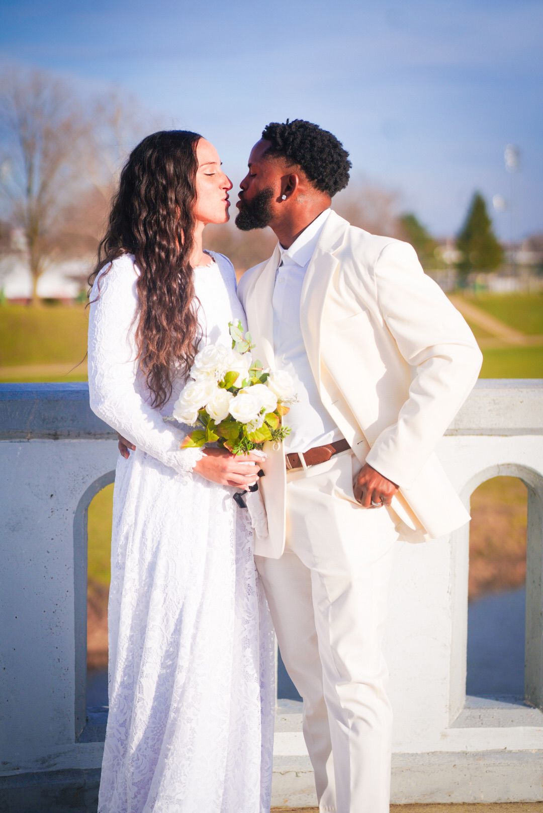 A couple shares romantic moments by a waterfront railing in white attire.