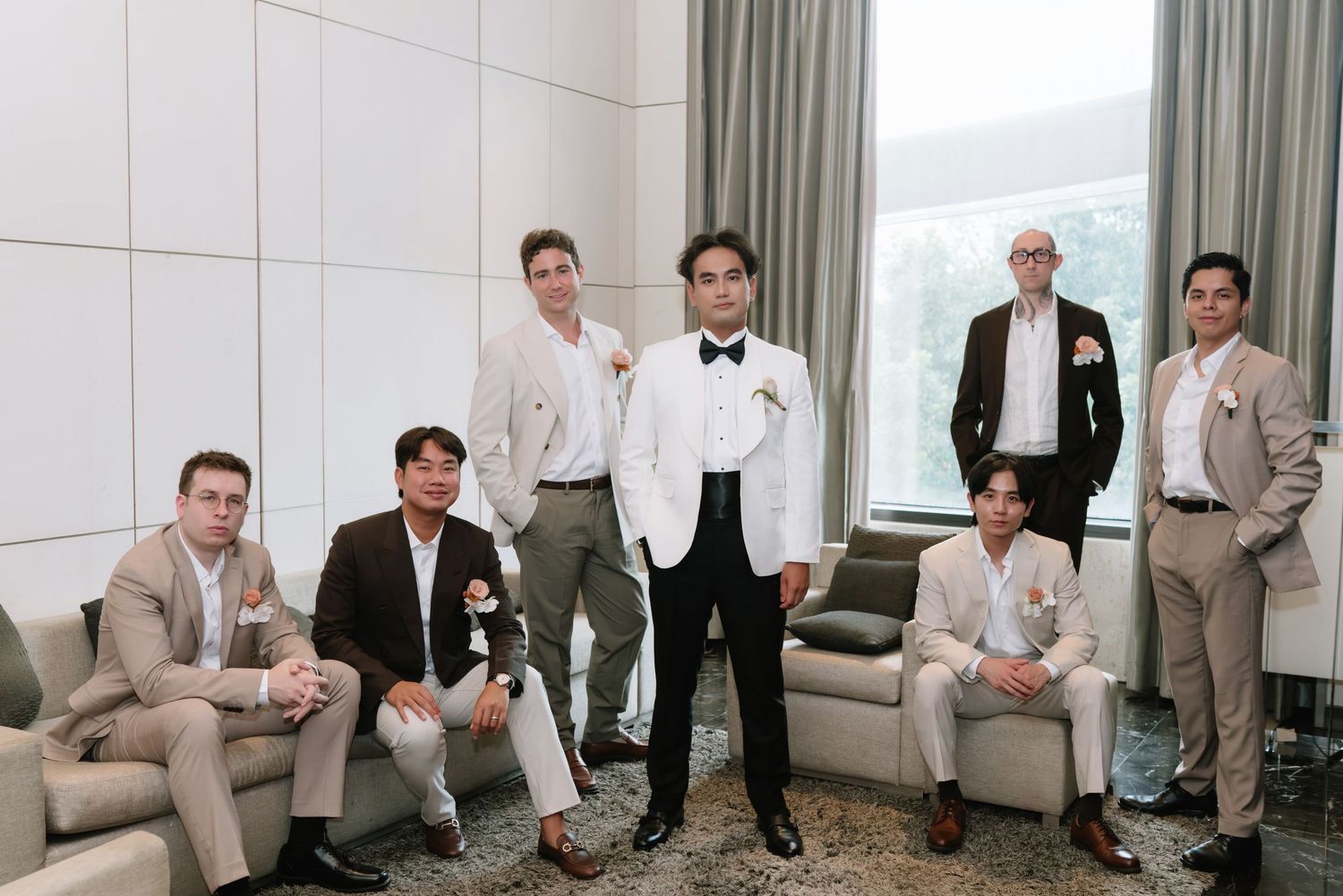 Groom in tuxedo poses with groomsmen in neutral suits in a modern indoor setting before the wedding ceremony.